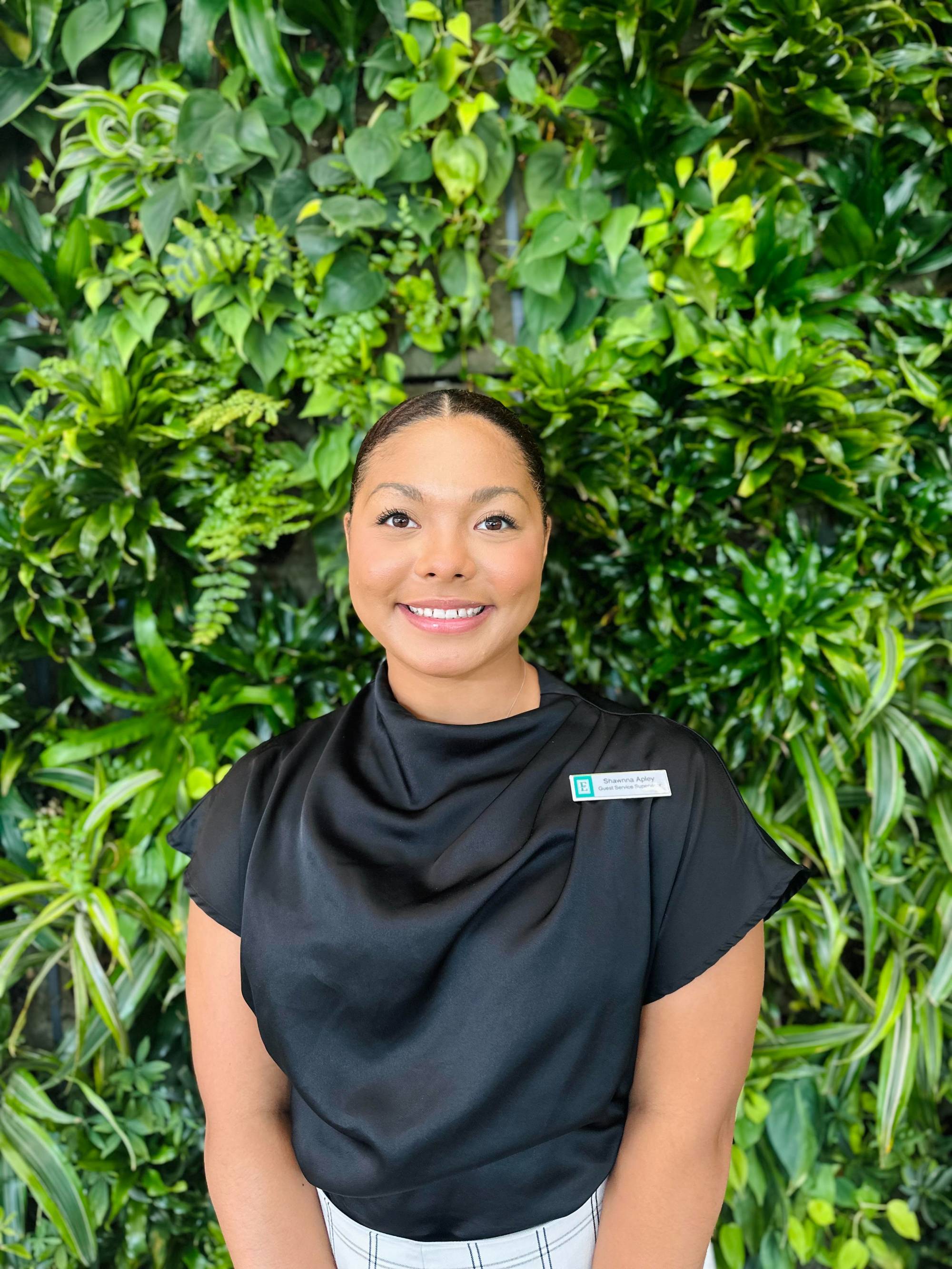 African American woman in a black shirt standing in front of a wall of plants